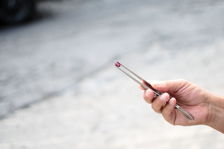 Hand holding a small red gemstone in tweezers for close scrutiny, the careful look sophisticated synthetics demand