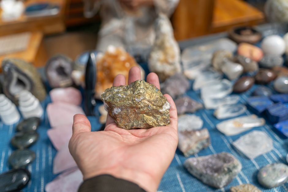 Gem Shows Are Worth It — Examine the Stone Before You Buy Hand examining a raw pyrite chunk over a gem show stall stocked with rose quartz, amethyst, and geodes