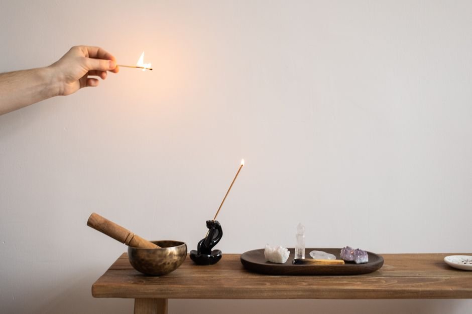 Hand lighting an incense stick beside a tray of amethyst and quartz stones to cleanse and recharge a crystal